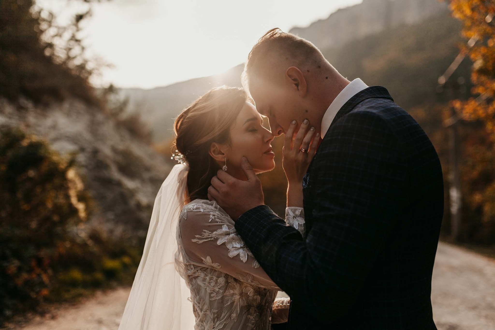 Para młoda w plenerze: panna młoda w koronkowej sukni i welonie, pan młody w garniturze. Delikatne światło słoneczne, subtelny makijaż, naturalne tło górskie.