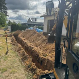 Wykopany wąski rów w ziemi, prawdopodobnie pod instalacje, widoczny z perspektywy kabiny koparki. W tle budynek w trakcie budowy i pracownik.