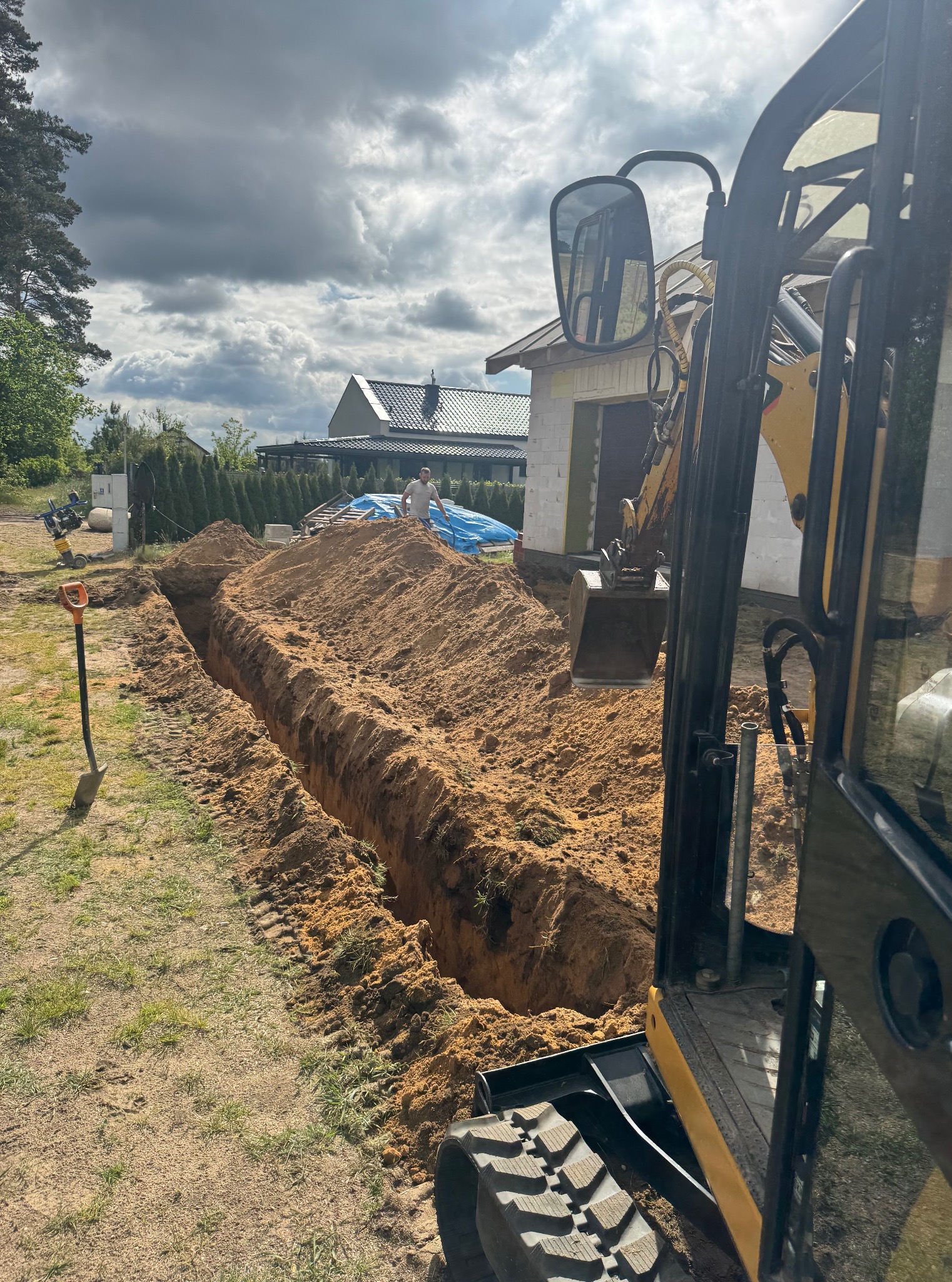Wykopany wąski rów w ziemi, prawdopodobnie pod instalacje, widoczny z perspektywy kabiny koparki. W tle budynek w trakcie budowy i pracownik.