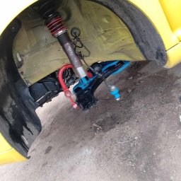 Clean Ride Serwis - Żółty samochód na podnośniku z widocznym, zdemontowanym zawieszeniem, sprężyną i amortyzatorem. Elementy zawieszenia w kolorze niebieskim i czerwonym.