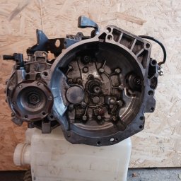 Clean Ride Serwis - Otwarta skrzynia biegów, widoczne elementy wewnętrzne, osadzona na białym zbiorniku, na tle płyty OSB i opony. Elementy mechaniczne pokryte olejem.