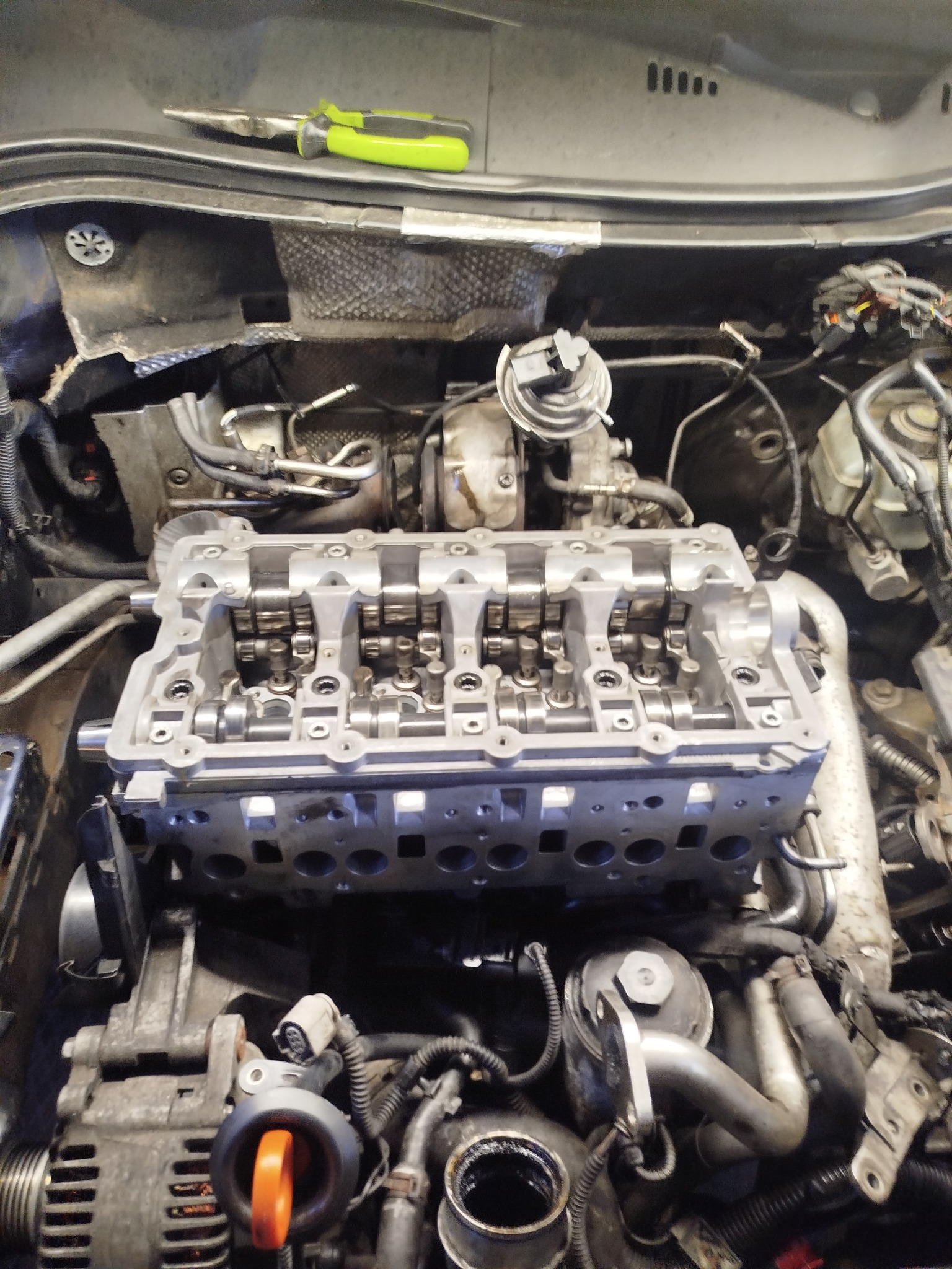 Silnik samochodowy z otwartą głowicą cylindrów, widoczne wałki rozrządu i elementy układu smarowania. Na górze leżą szczypce z zieloną rękojeścią.