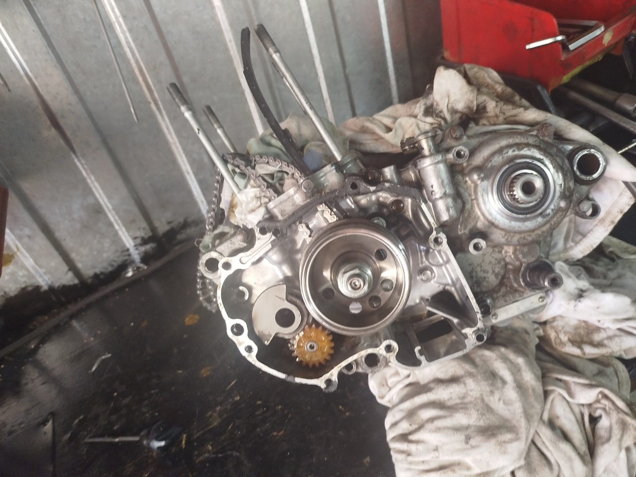 Rozłożony silnik motocyklowy na warsztatowym stole, widoczne tłoki, łańcuch, przekładnia, elementy mocowania i brudne szmaty. Częściowo widoczna skrzynka z narzędziami.