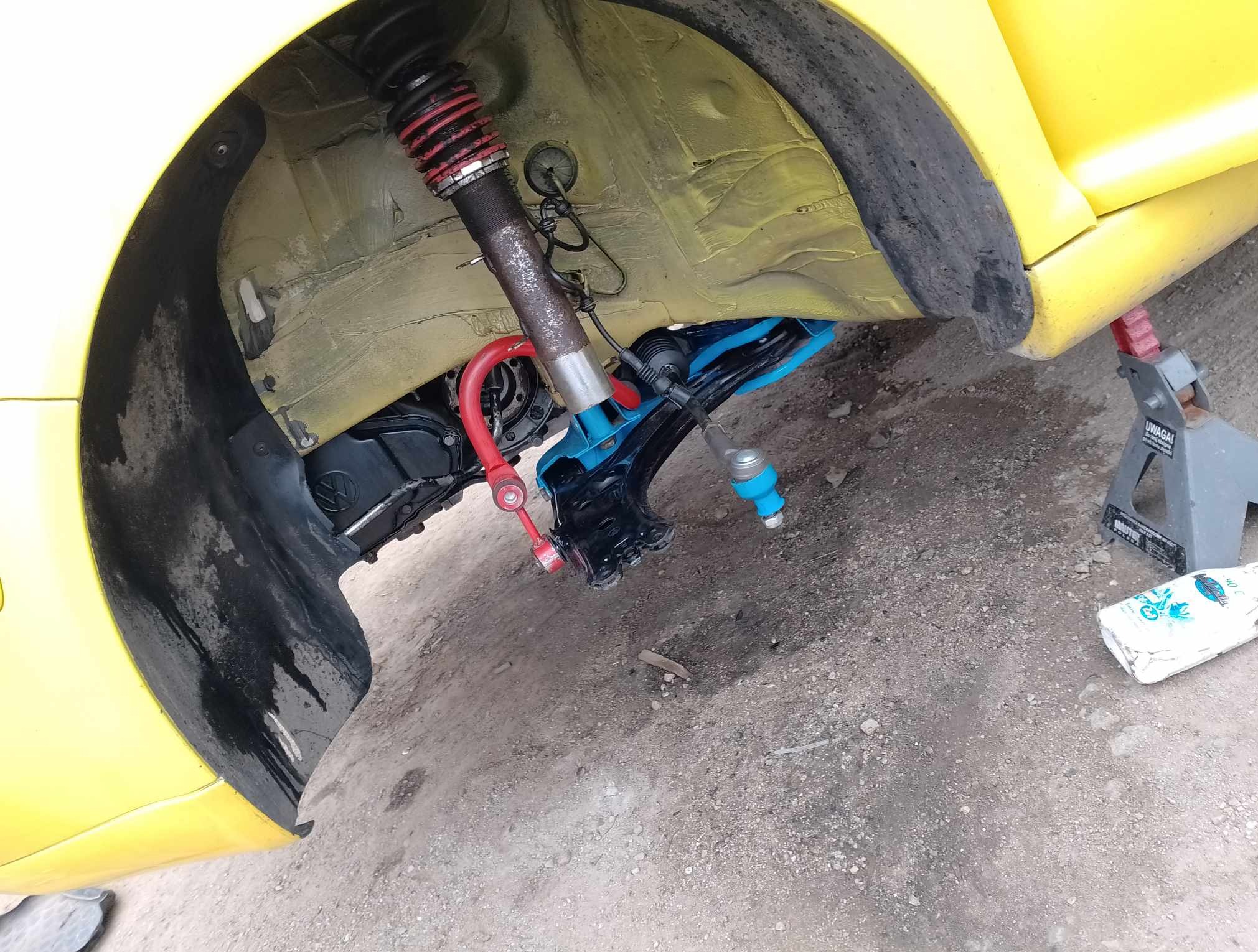 Żółty samochód na podnośniku z widocznym, zdemontowanym zawieszeniem, sprężyną i amortyzatorem. Elementy zawieszenia w kolorze niebieskim i czerwonym.