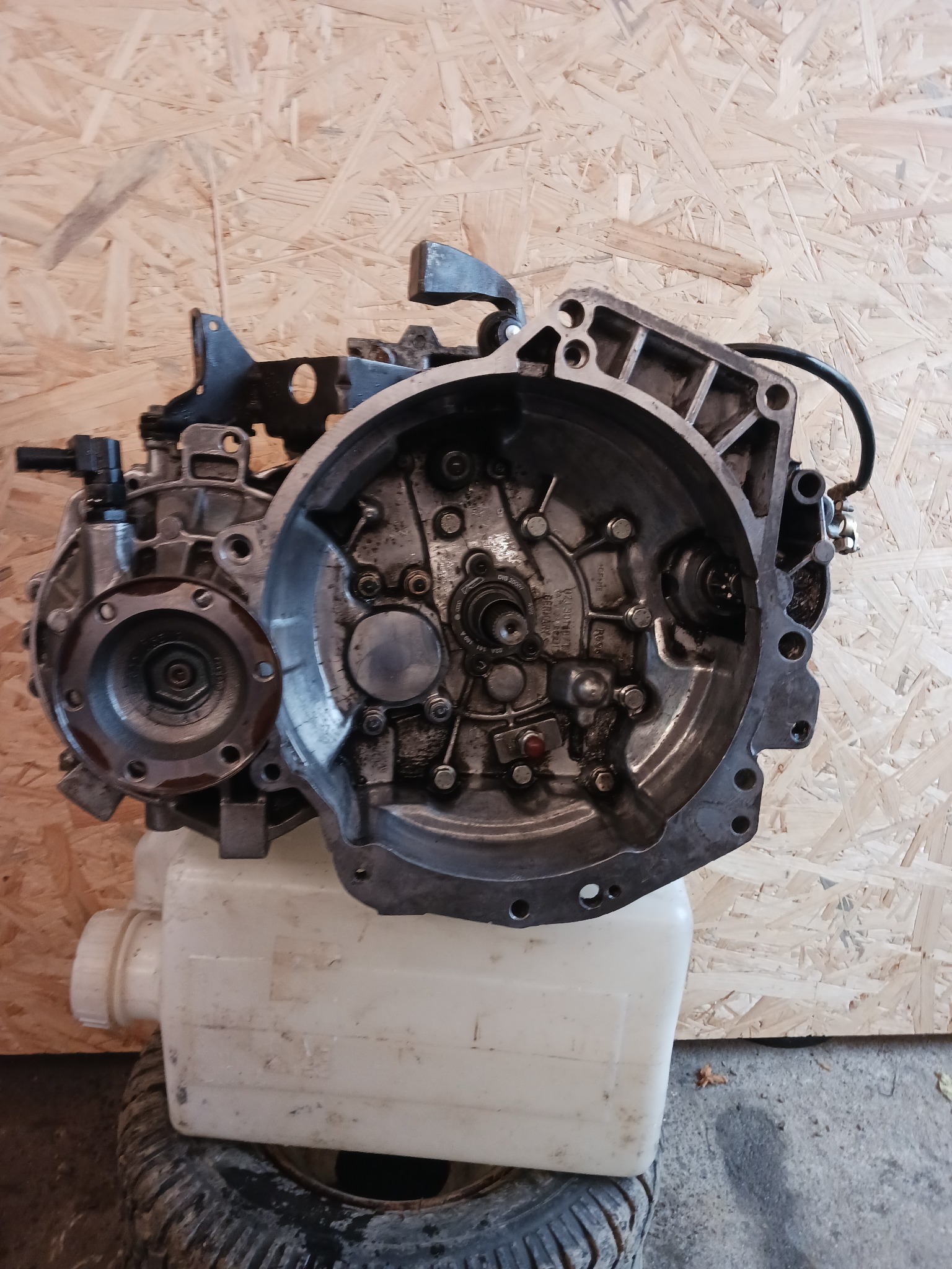 Otwarta skrzynia biegów, widoczne elementy wewnętrzne, osadzona na białym zbiorniku, na tle płyty OSB i opony. Elementy mechaniczne pokryte olejem.