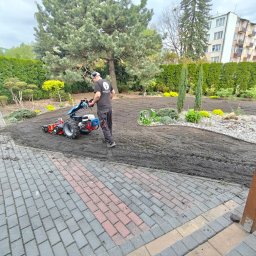 Botanicus Usługi Ogrodnicze Bartłomiej Korzeniowski - Mężczyzna z glebogryzarką spalinową przygotowuje ziemię pod trawnik w ogrodzie. W tle drzewa, krzewy i budynek mieszkalny. Obrzeże z kostki brukowej.