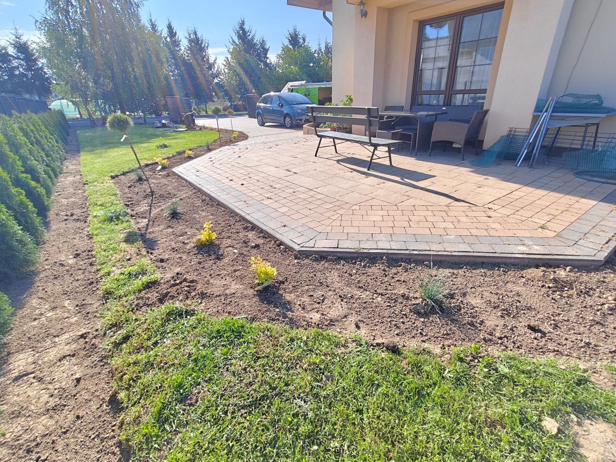 Układanie kostki brukowej na tarasie, świeżo posadzone rośliny wzdłuż ścieżki, schludny trawnik i zadbane tuje w słoneczny dzień. Widok na dom i podjazd.