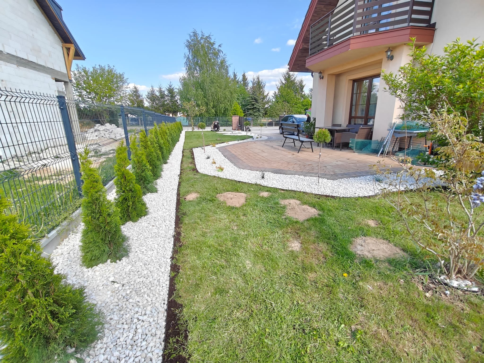 Aranżacja ogrodu z tujami wzdłuż ogrodzenia i białymi kamieniami, trawnik z kamiennymi ścieżkami, patio z meblami ogrodowymi, dom z balkonem.