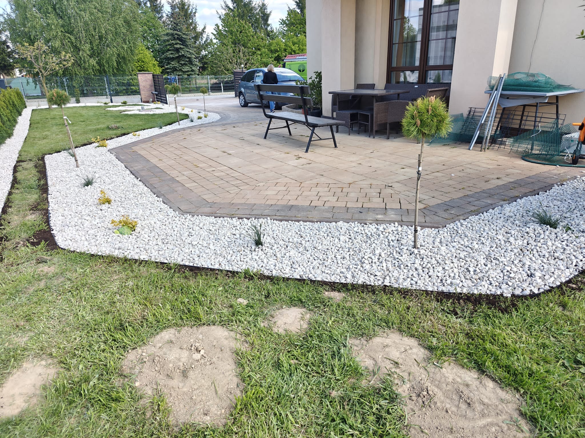 Wykończony fragment ogrodu z wybrukowanym patio, obłożonym białym kamieniem i nasadzeniami. Widoczna ławka, meble ogrodowe i drzewka kuliste. Osoba przy samochodzie w tle.