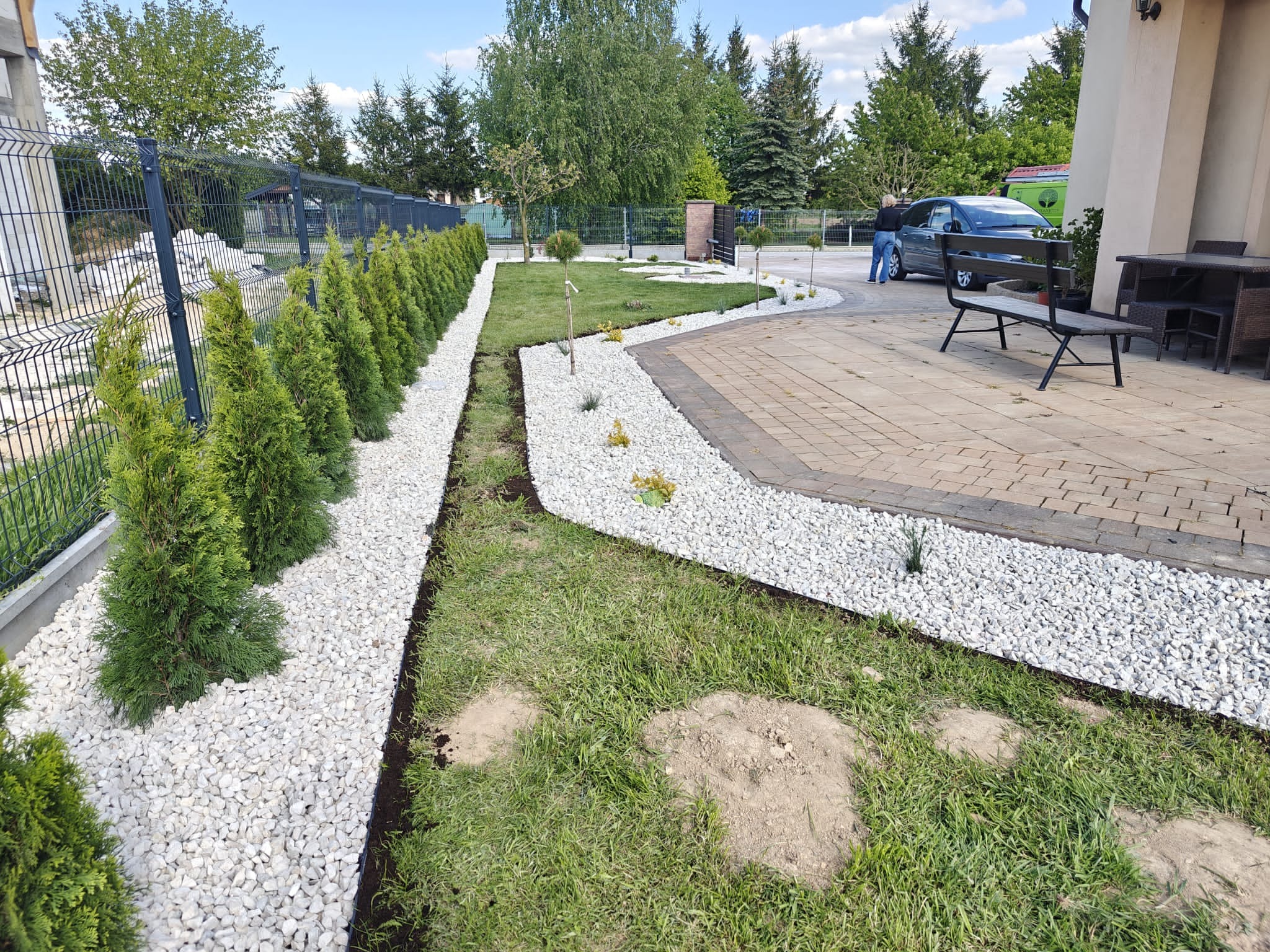 Ogród po metamorfozie: rząd tuj wzdłuż siatki, białe kamienie oddzielające roślinność od trawnika, kostka brukowa, drzewa w tle i osoba przy samochodzie.
