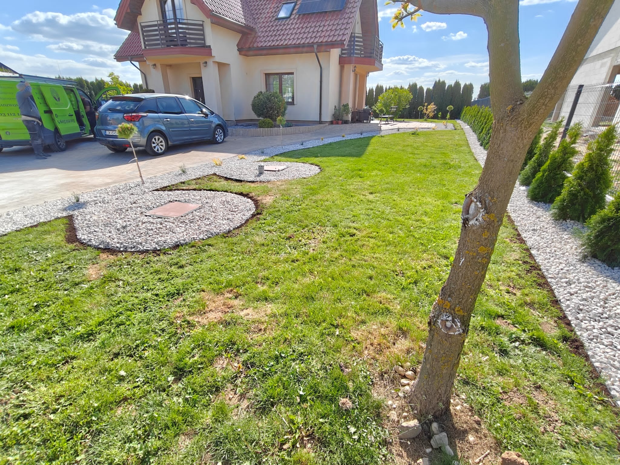 Ogród po realizacji: trawnik z nieregularnymi rabatami z białego żwiru, obrzeża z kamyków, tuje wzdłuż ogrodzenia, dom z balkonem i zaparkowany samochód.