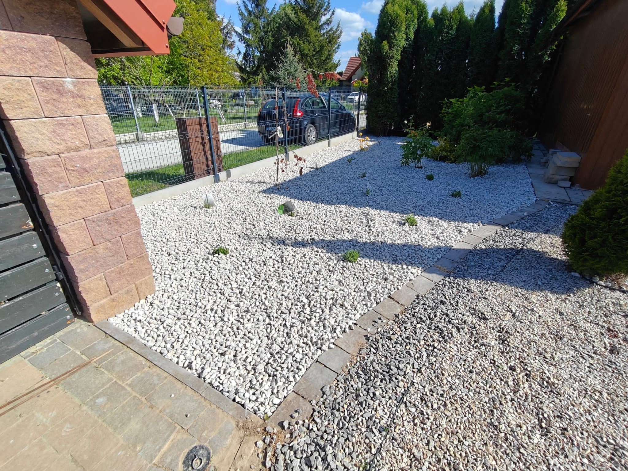 Ogród z białym żwirem i obrzeżami z kostki, z zielonymi krzewami i drzewami w tle. Widoczny fragment domu z cegły i metalowe ogrodzenie.