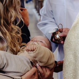 Usługi Kozłowski - Chrzest dziecka: ksiądz polewa wodą główkę niemowlęcia trzymanego przez matkę w beżowym sweterku. W tle widoczni goście i elementy wystroju kościoła.