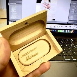 Usługi Kozłowski - Pamiątka Chrztu Świętego: drewniany pendrive z grawerem imienia 'Maksia' w otwartym pudełku, na tle laptopa z otwartym zdjęciem dziecka po obróbce.