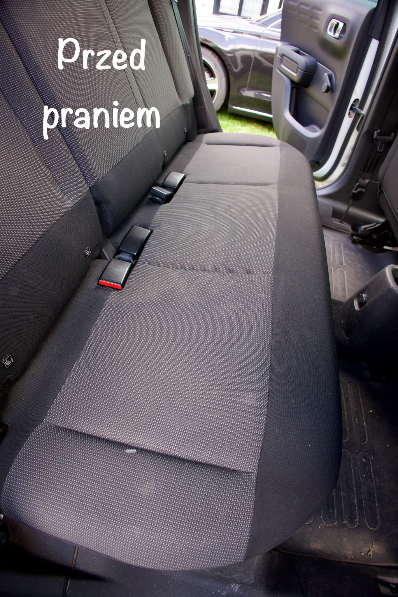 Szara, materiałowa tapicerka tylnej kanapy samochodowej przed praniem, z widocznymi pasami bezpieczeństwa i elementami wnętrza pojazdu.