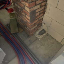 Waszka - Fragment ściany z czerwonej cegły i białych bloczków, z widocznymi rurami instalacji. Otwór uszczelniony pianką montażową. Detal prac wykończeniowych.