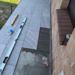 Waszka - Układanie szarych płytek na balkonie przy ceglanej ścianie. Widoczne poziomice, krzyżyki dystansowe i kielnia. Częściowo położone płytki z widocznym klejem.