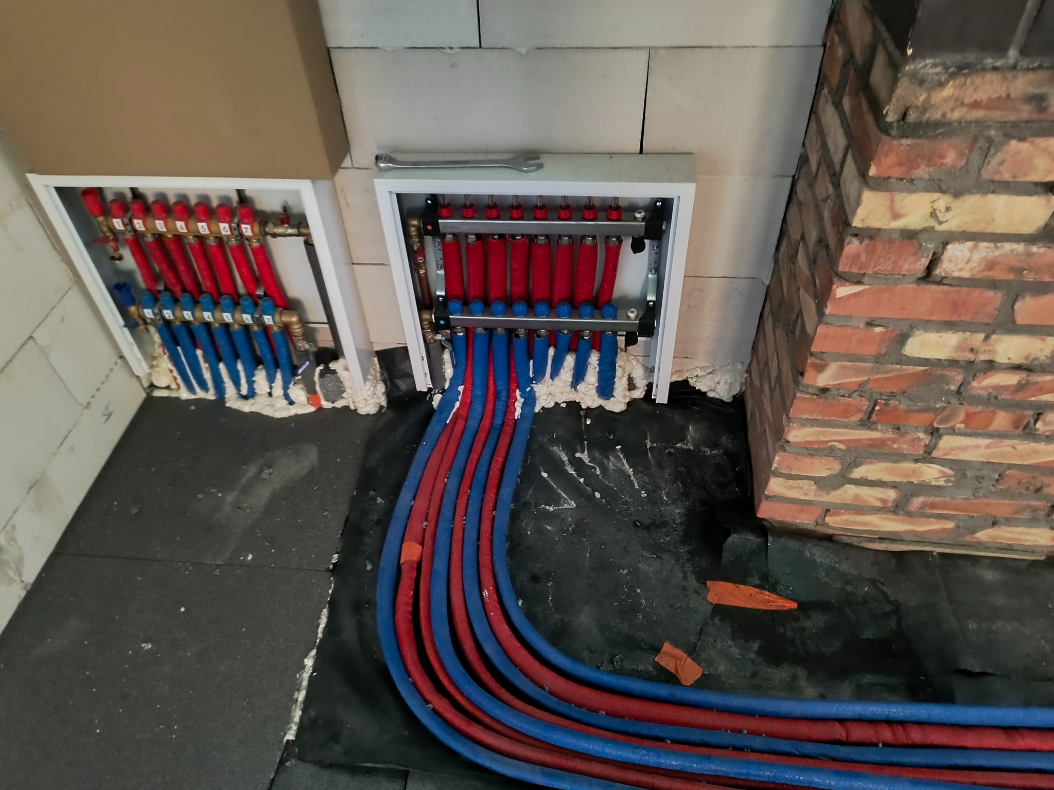 Rozdzielacz ogrzewania podłogowego z czerwono-niebieskimi rurami, zamontowany w białej skrzynce, widoczny klucz płaski, ceglana ściana w tle, czarna izolacja podłogowa.