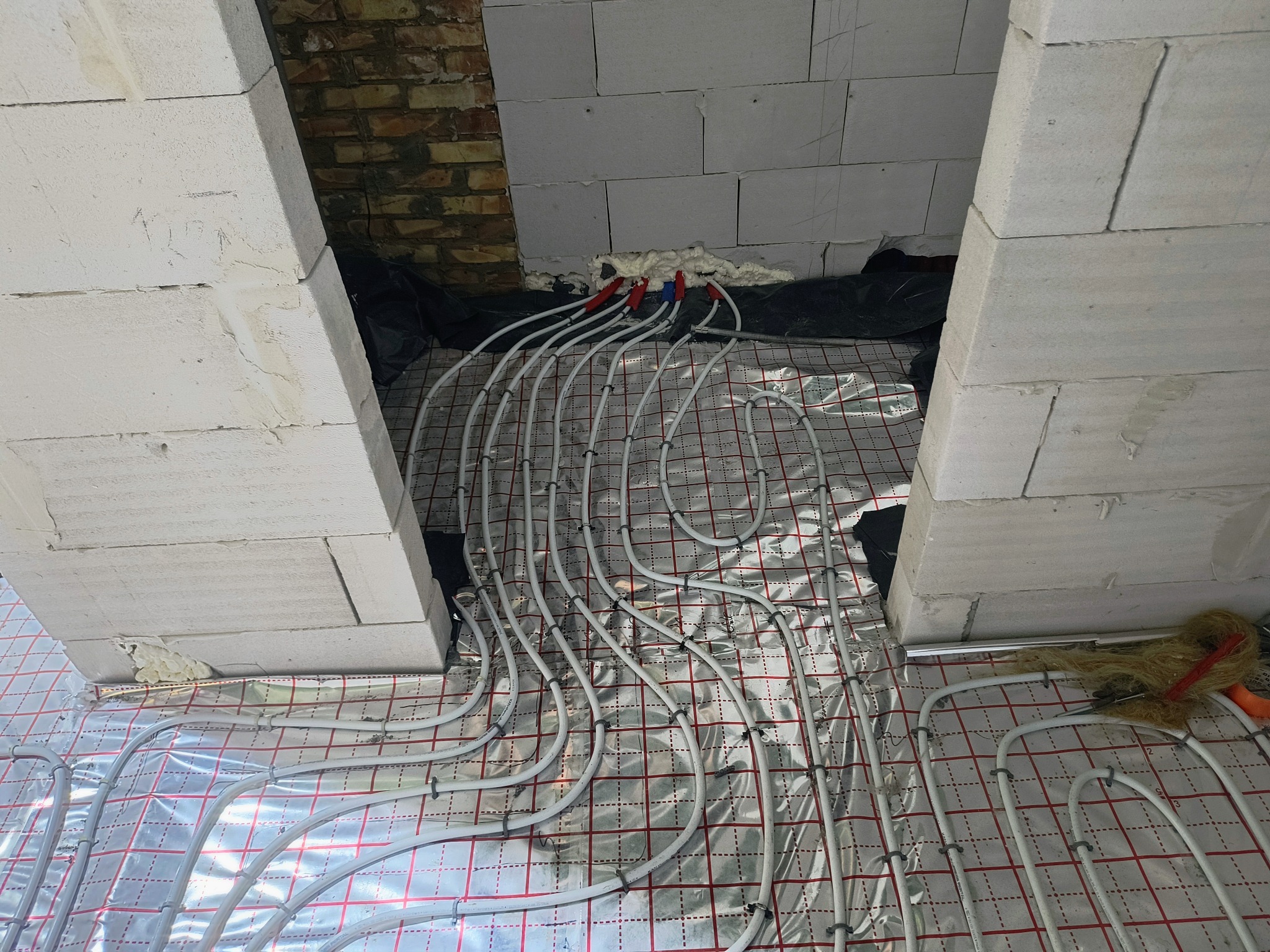 Instalacja ogrzewania podłogowego w trakcie montażu, rury ułożone na folii z siatką, widoczne ściany z bloczków i fragment instalacji hydraulicznej.