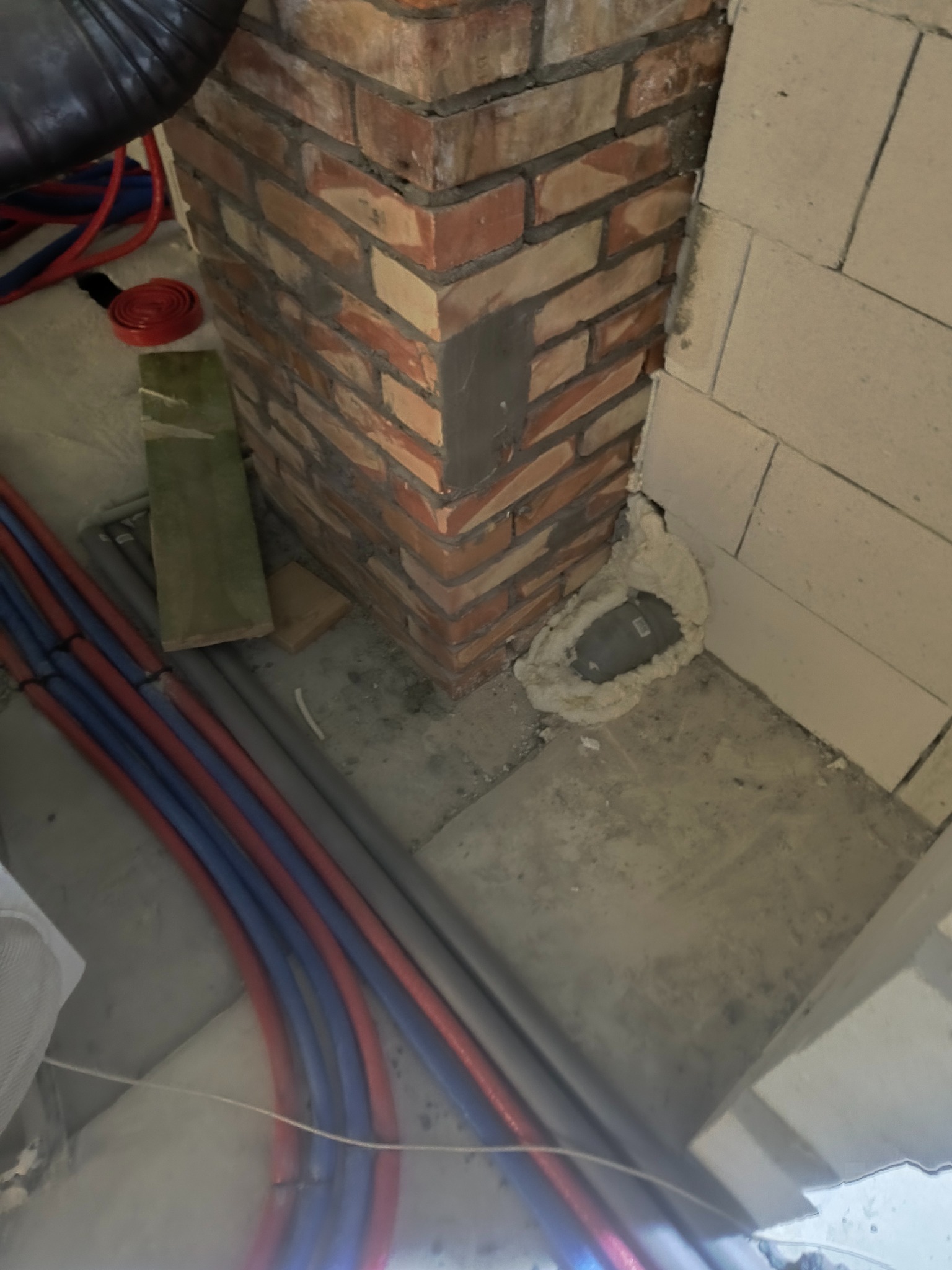 Fragment ściany z czerwonej cegły i białych bloczków, z widocznymi rurami instalacji. Otwór uszczelniony pianką montażową. Detal prac wykończeniowych.