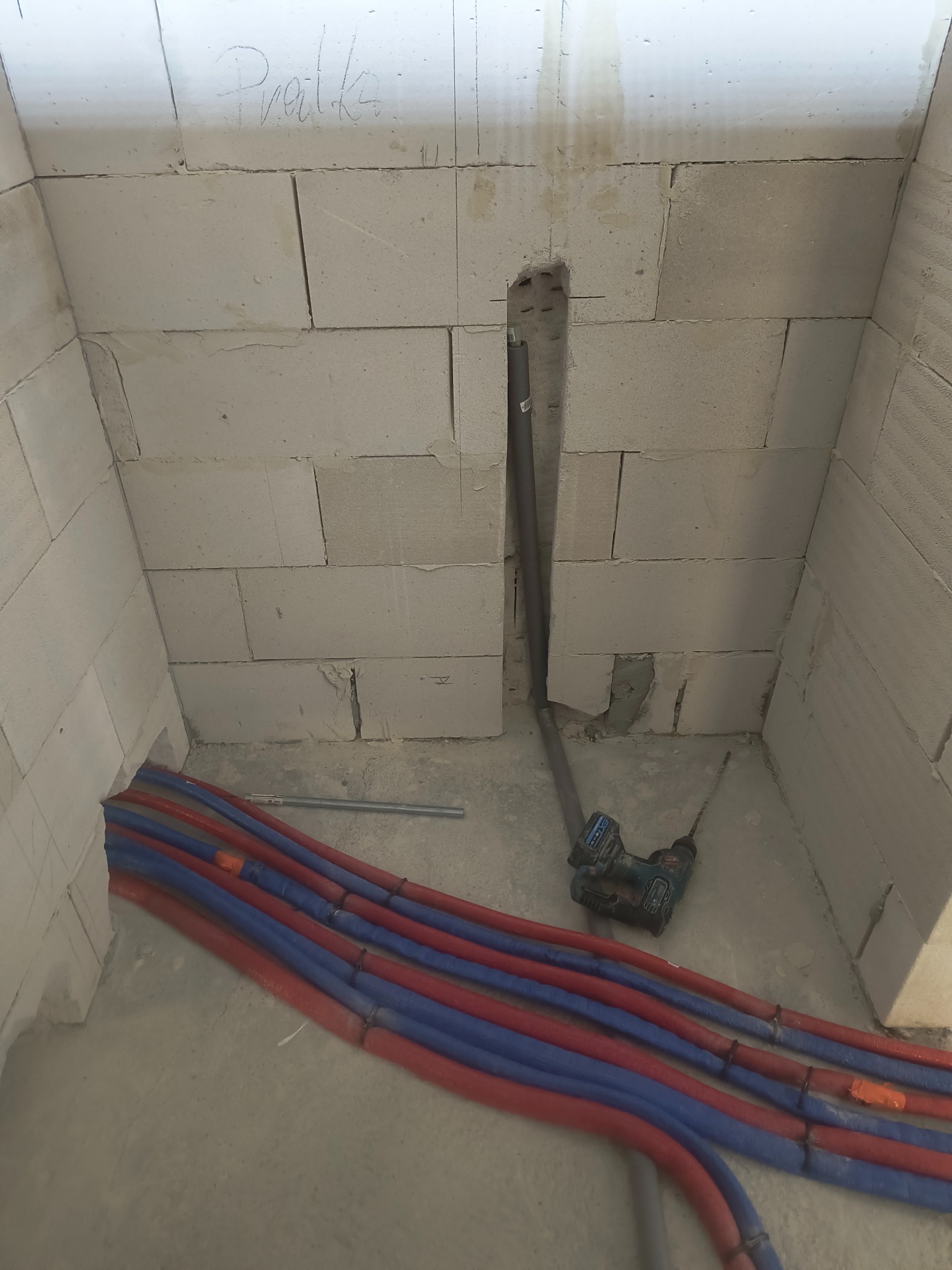Instalacja wodna w trakcie montażu: rury PEX w kolorach czerwonym i niebieskim ułożone na podłodze, w tle ściana z bloczków betonowych z wywierconym otworem na rury.