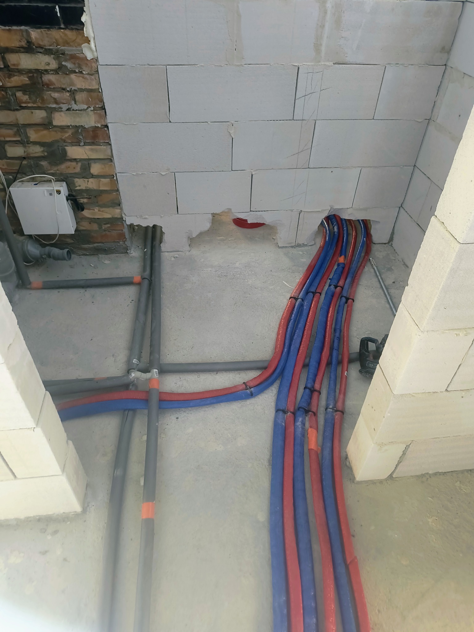 Instalacja hydrauliczna w trakcie montażu: czerwono-niebieskie rury prowadzone w ścianie z białych bloczków, szare rury łączone złączkami. Widoczny fragment ściany z cegły.