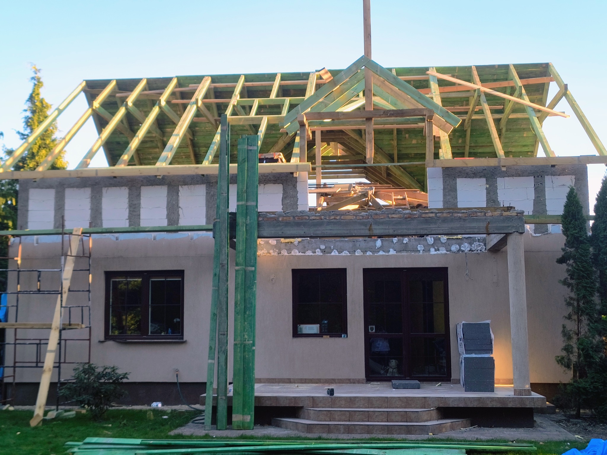 Rozbudowa domu jednorodzinnego: widok na konstrukcję dachu z drewnianych belek, częściowo wymurowane ściany z bloczków i rusztowanie. Etap budowy w słoneczny dzień.