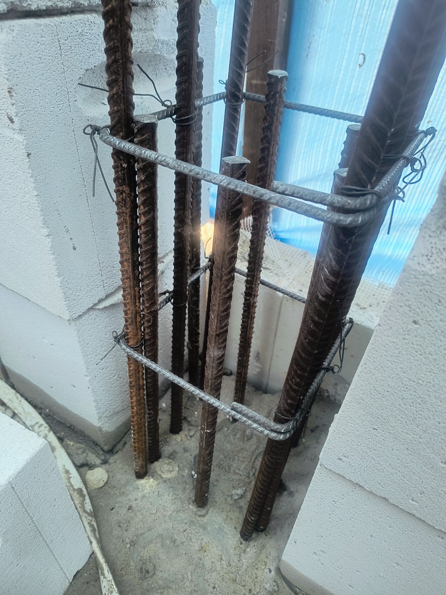 Zbrojenie pionowe z żebrowanych prętów stalowych, połączonych drutem, osadzone w betonie, przy białych bloczkach. Widoczna niebieska folia w tle.