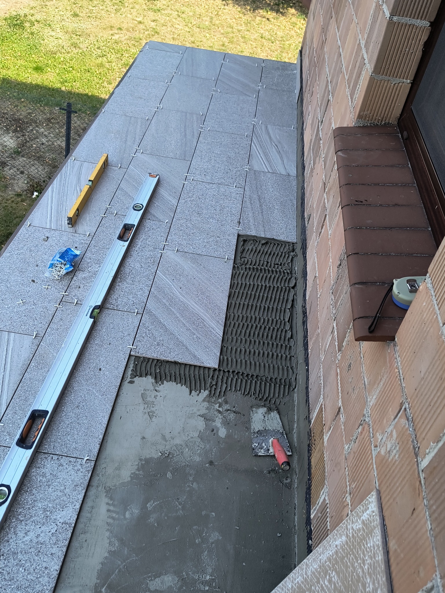 Układanie szarych płytek na balkonie przy ceglanej ścianie. Widoczne poziomice, krzyżyki dystansowe i kielnia. Częściowo położone płytki z widocznym klejem.