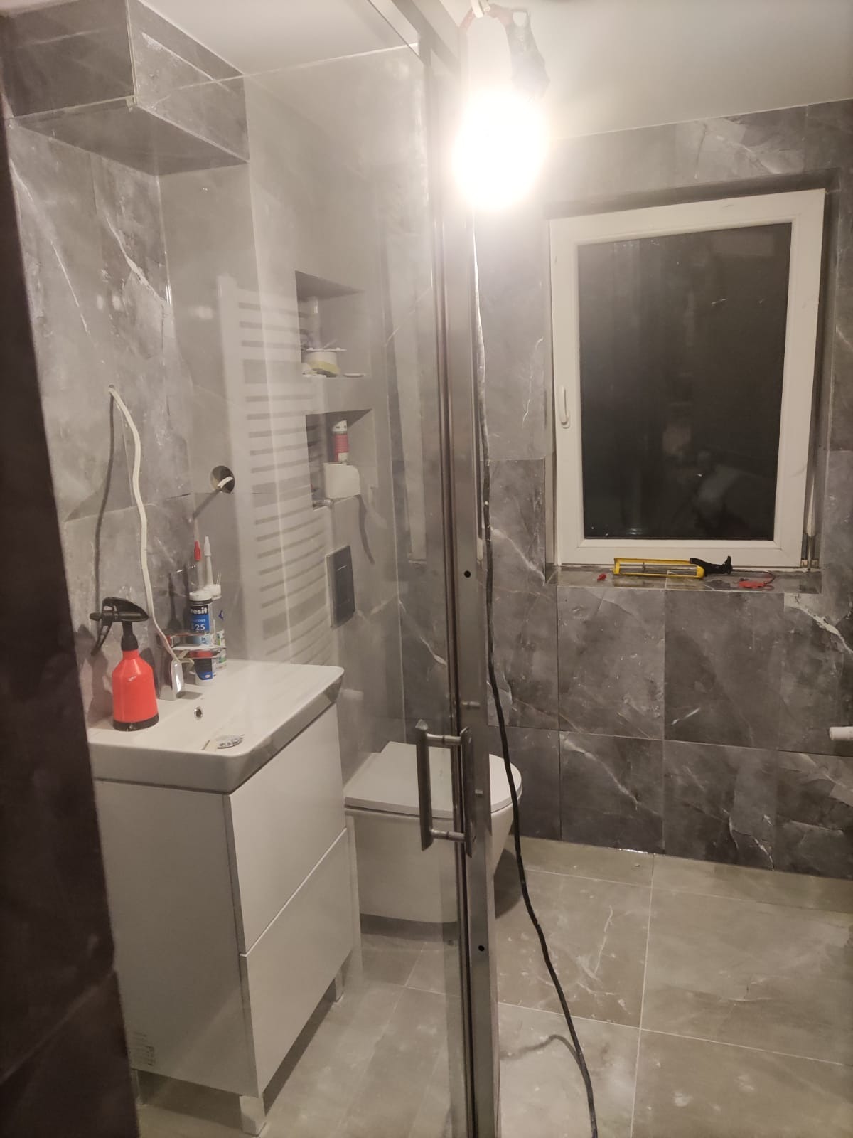 Wykończona łazienka z szarymi płytkami imitującymi marmur, białą armaturą i kabiną prysznicową. Widoczne narzędzia i akcesoria, sugerujące świeżo zakończony remont.