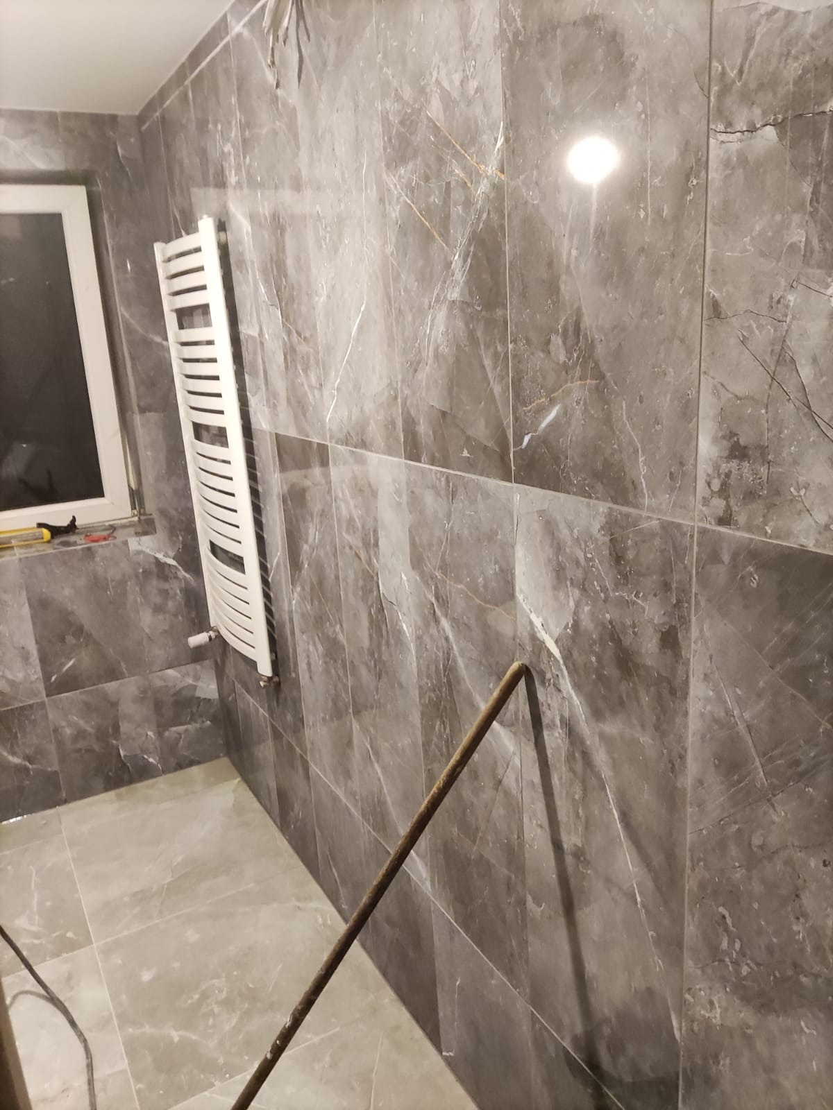 Wykończona łazienka z szarymi płytkami imitującymi marmur, grzejnikiem drabinkowym i widocznym narzędziem. Ujęcie z dołu, podkreślające wzór płytek.