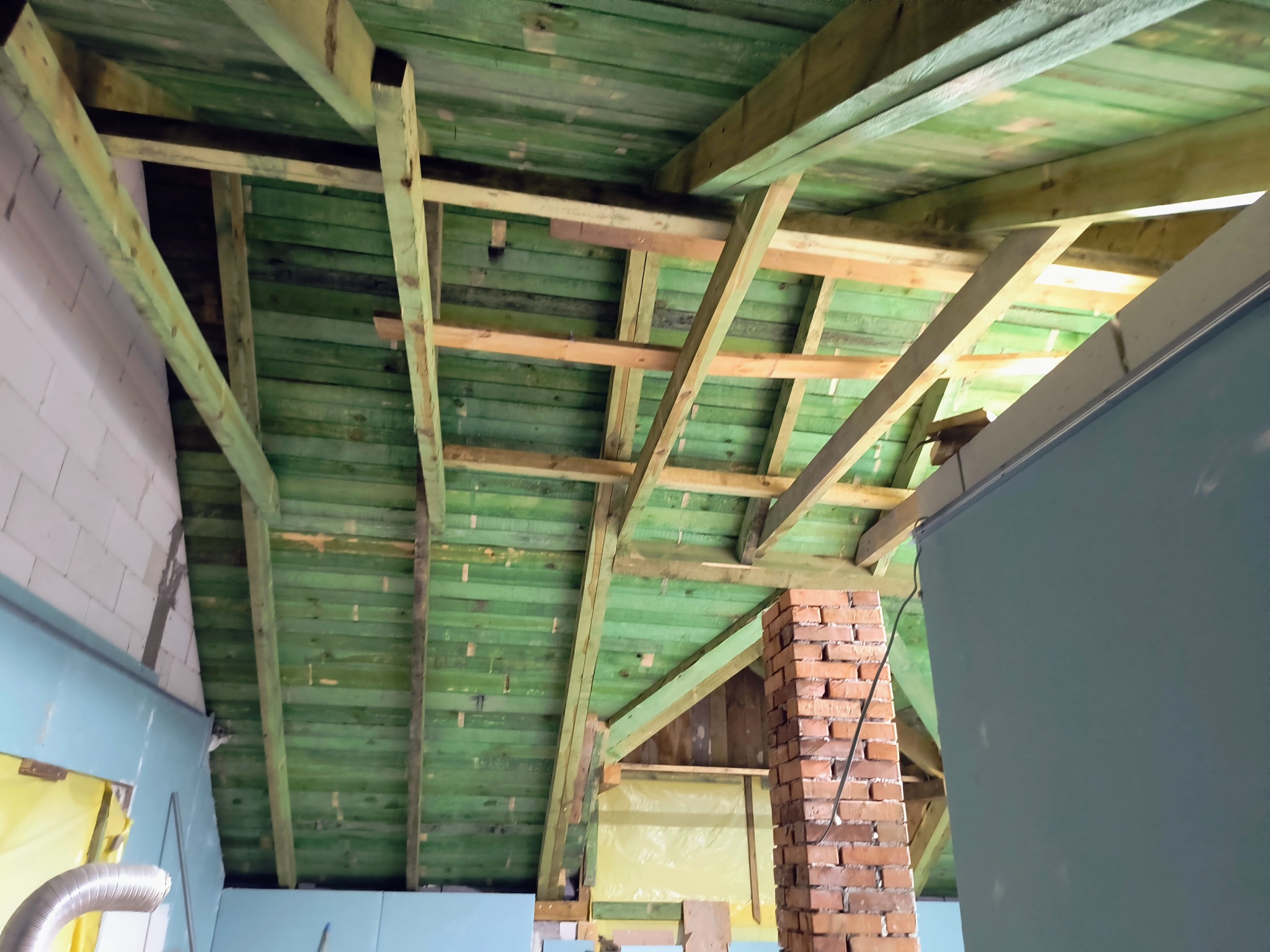 Wnętrze poddasza z widoczną konstrukcją dachu z impregnowanego drewna, fragmentem komina z cegły i ścianami w trakcie wykończenia płytami gipsowo-kartonowymi.