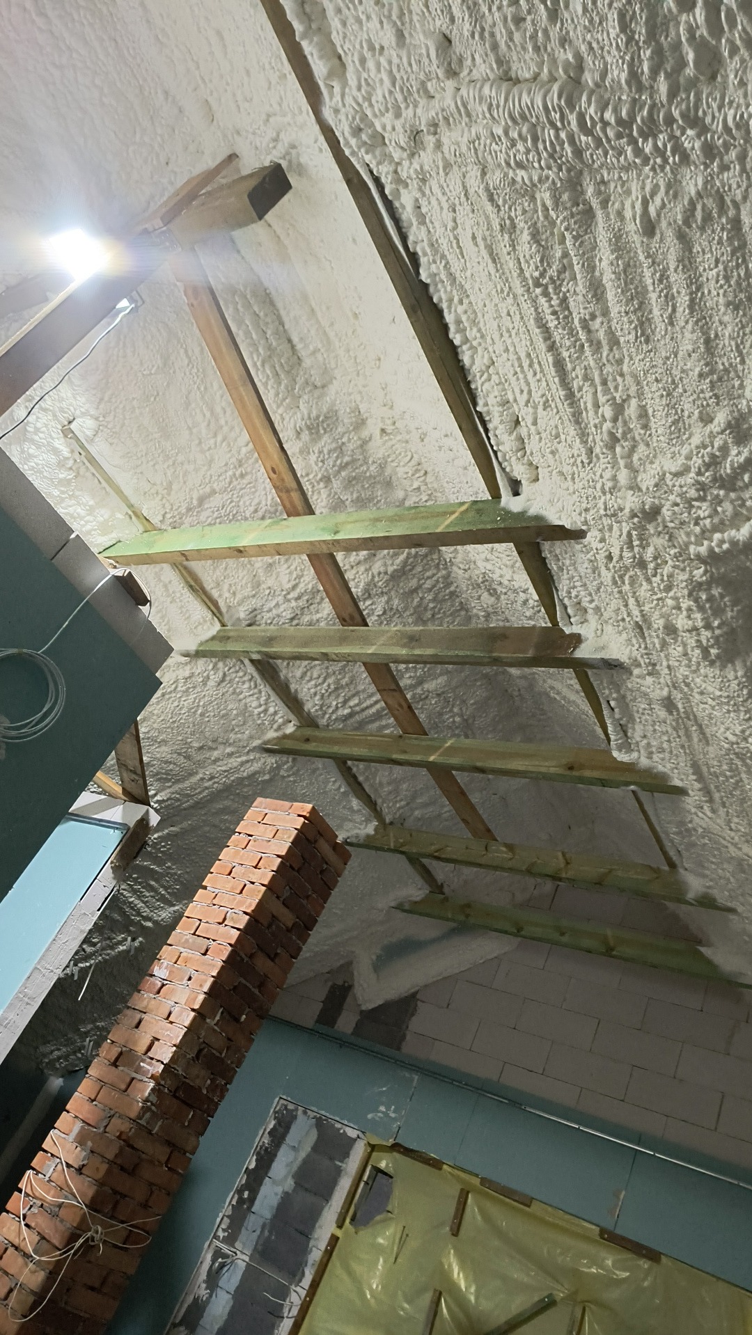 Poddasze z drewnianą konstrukcją i pianą izolacyjną, widok z dołu. Fragment ściany z cegły i płyty karton-gips. Oświetlenie punktowe.