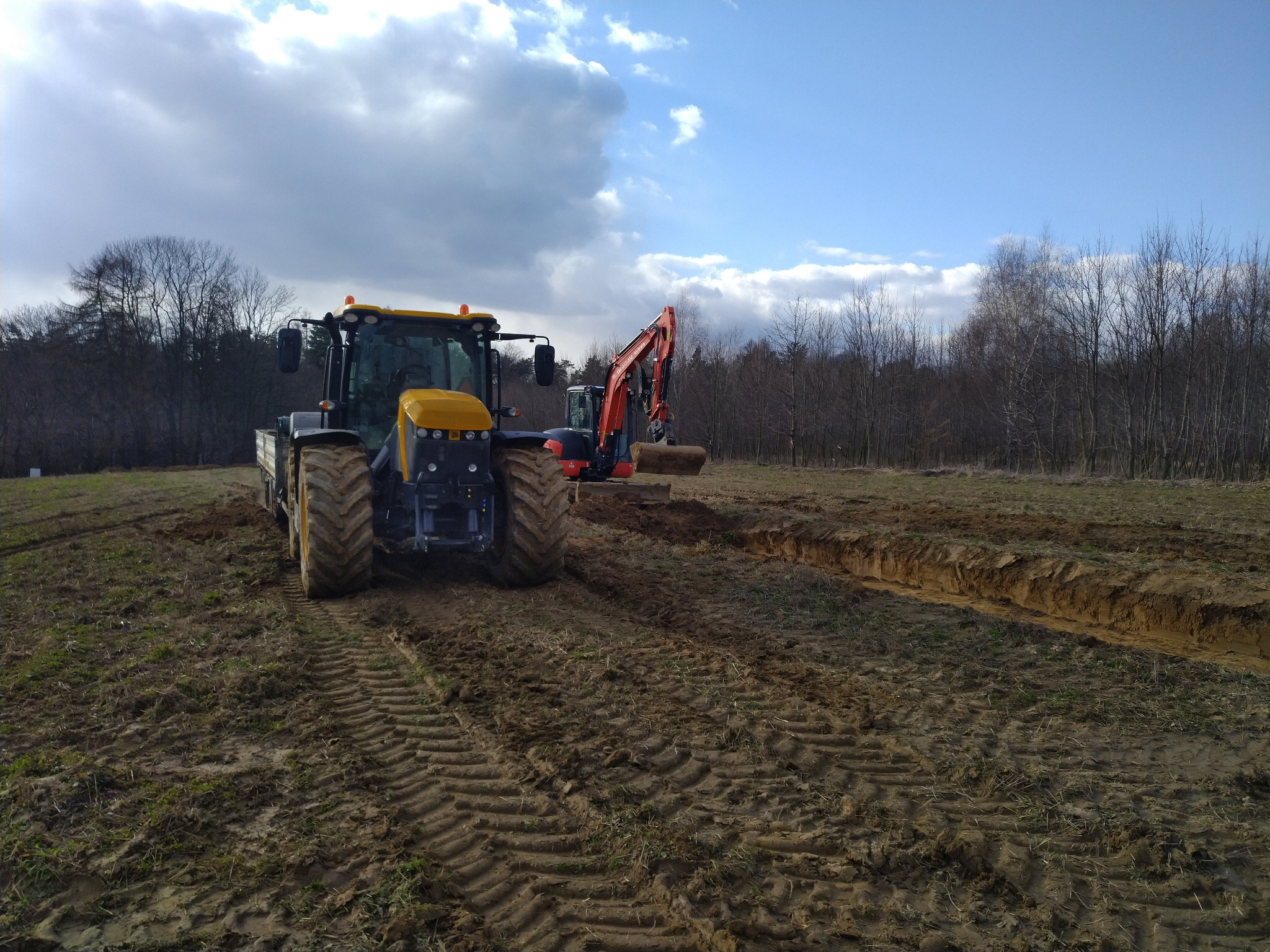 Żółty traktor i pomarańczowa koparka pracują na polu, tworząc rów. Ślady opon na ziemi, w tle drzewa i pochmurne niebo.