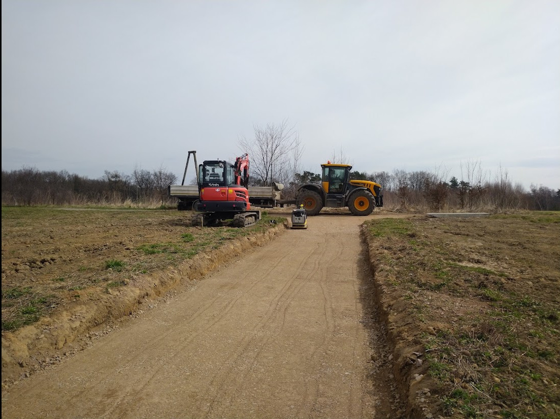 Żółty traktor i czerwona mini koparka z ubijarką na nowo utwardzonej, gruntowej drodze. W tle drzewa i pochmurne niebo. Widoczne ślady opon i pracy maszyn.