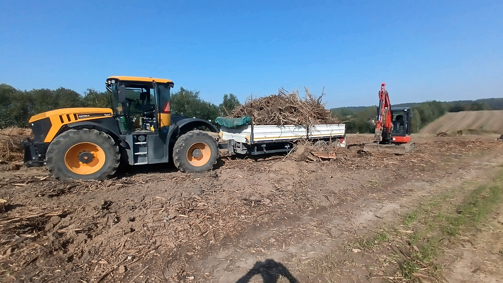 Żółto-czarny traktor z przyczepą pełną korzeni i gałęzi na polu po wycince, obok czerwona koparka. Błękitne niebo w tle. Widoczne ślady pracy maszyn.