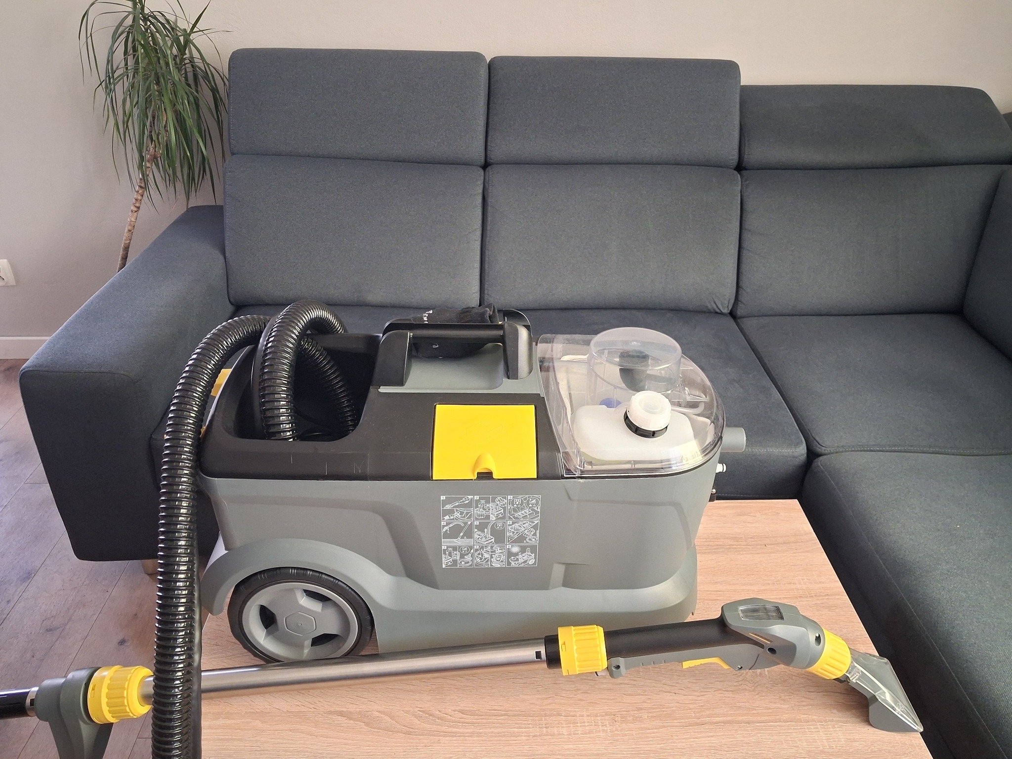 Szaro-żółty odkurzacz piorący z przezroczystym zbiornikiem na wodę, wąż i końcówka, stoi na stole obok szarej sofy. W tle widoczna roślina doniczkowa.