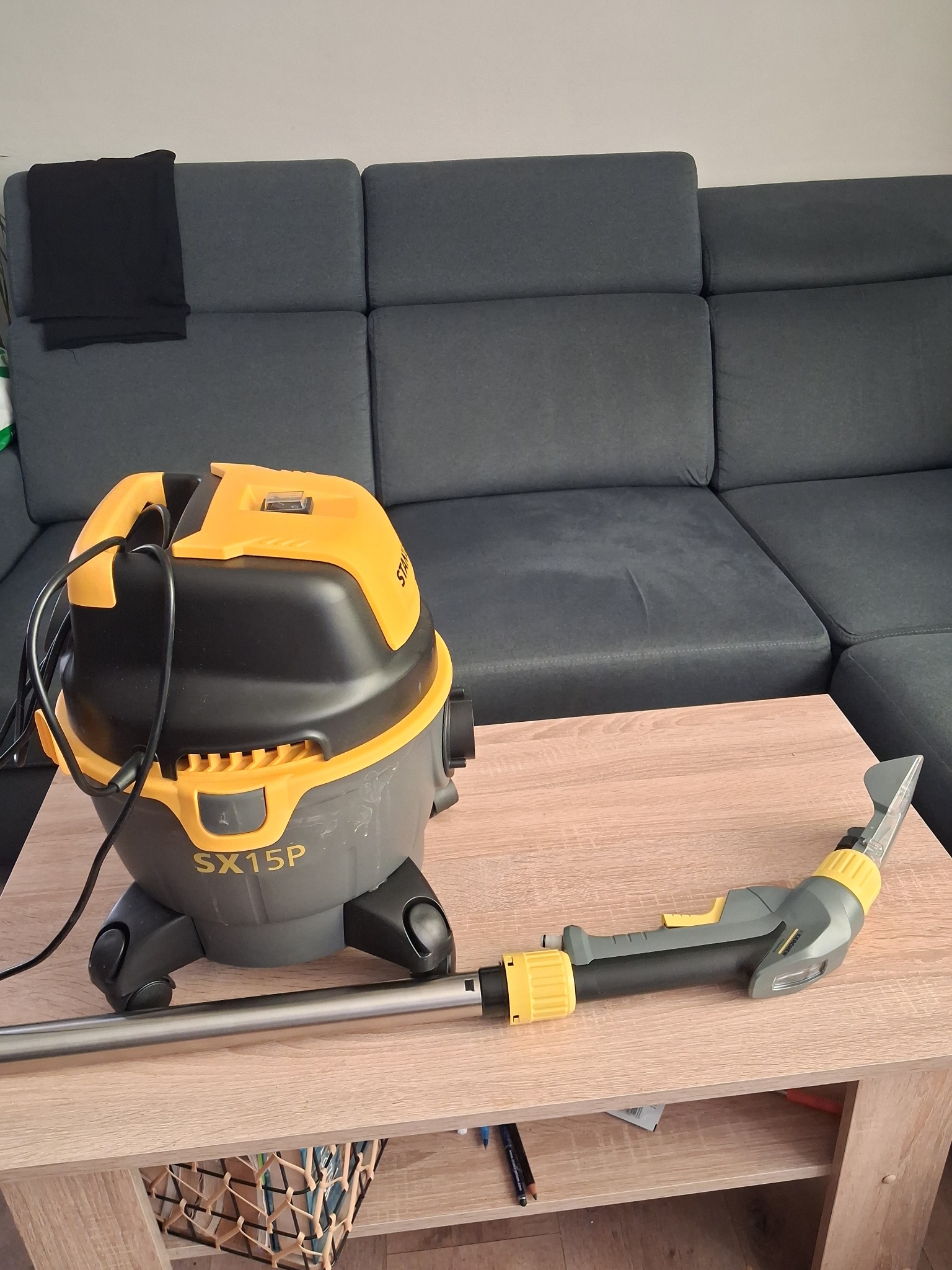 Żółto-szary odkurzacz piorący STANLEY SX15P z podłączoną końcówką do prania tapicerki, ustawiony na drewnianym stoliku, w tle szara kanapa.