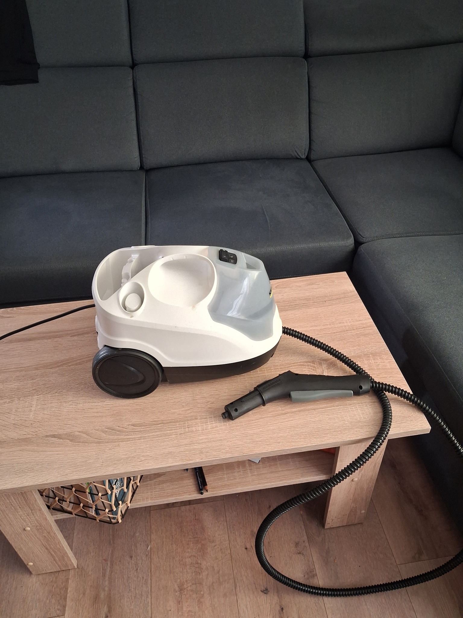 Parownica Karcher na stoliku kawowym obok szarej sofy. Widoczny wąż i końcówka parowa. Sprzęt gotowy do czyszczenia tapicerki.