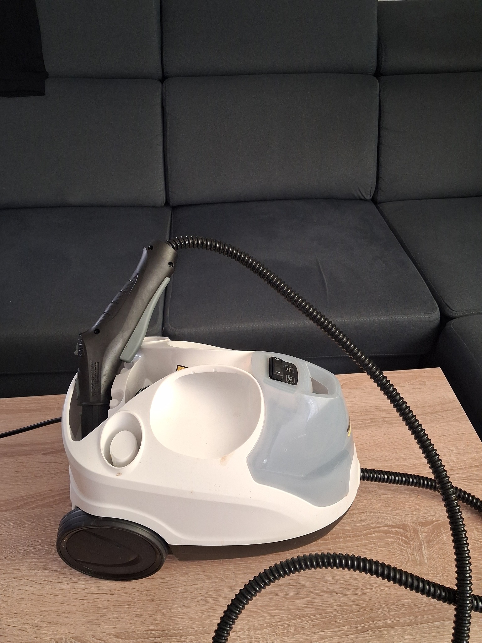 Parownica Karcher na drewnianym stoliku, gotowa do użycia. W tle widoczna szara sofa. Sprzęt czyszczący, czyszczenie parą, domowe porządki.