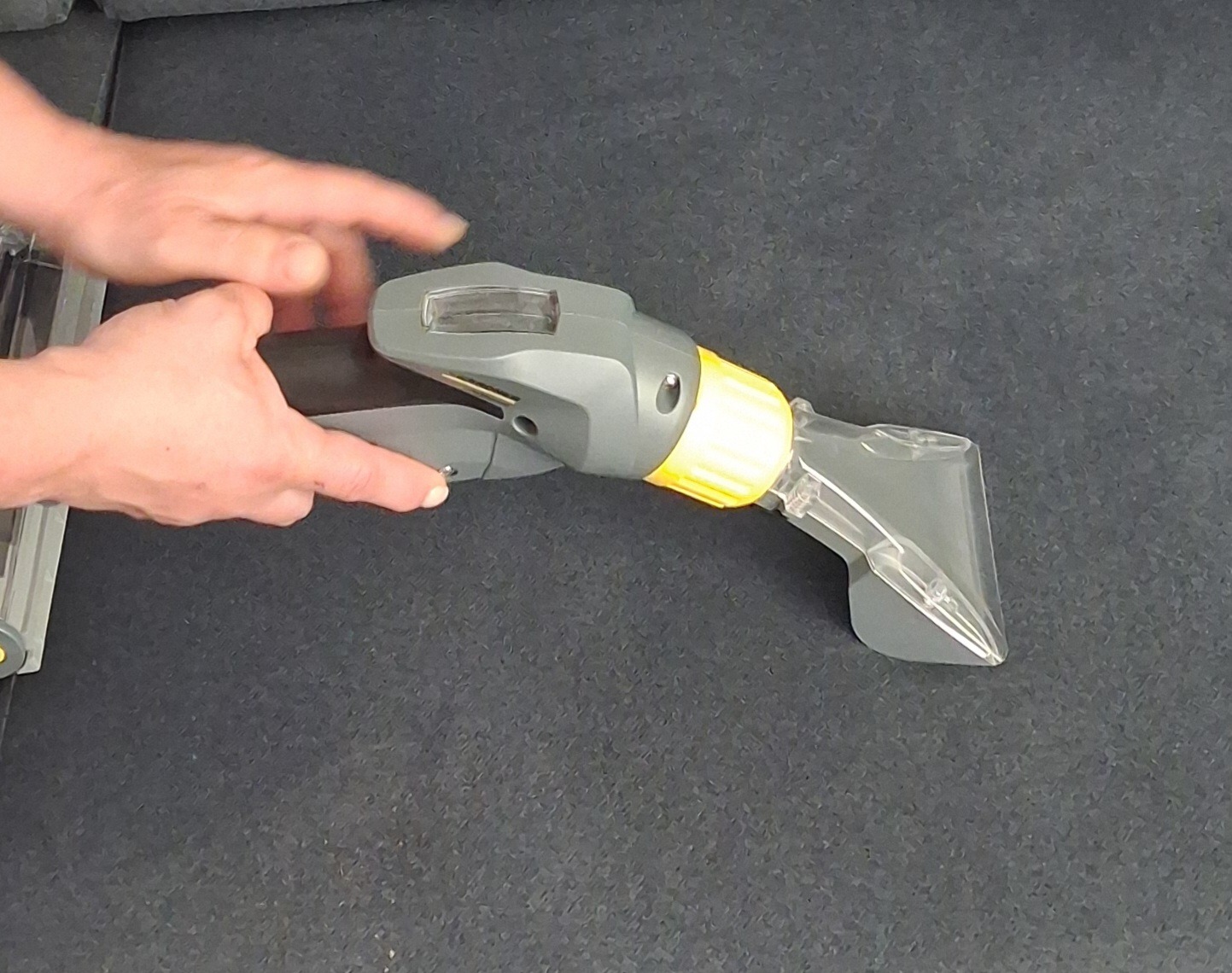 Dysza odkurzacza ekstrakcyjnego w trakcie czyszczenia ciemnej tapicerki. Widoczne dłonie operatora. Sprzęt w kolorze szarym z żółtymi elementami.