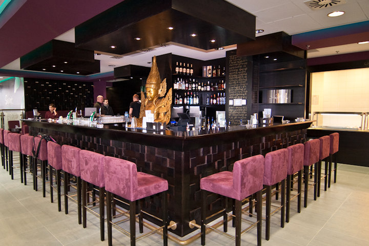 Wnętrze restauracji azjatyckiej SHANTI z długim, ciemnym barem, rzędem purpurowych krzeseł barowych, złotą statuetką Buddy i półkami z butelkami alkoholu.