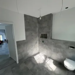 Łazienka w trakcie wykańczania: szare płytki, białe ściany, surowa żarówka. Widoczne niedokończone instalacje elektryczne i sanitarna. W tle otwarte drzwi.