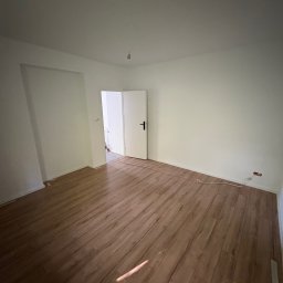 PRZEMYSŁAW DZIEDZIC PEPWO - Puste, jasne pomieszczenie z panelami podłogowymi w odcieniach brązu. Widoczne białe ściany, otwarte drzwi i wystające kable elektryczne przy podłodze.