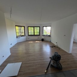 PRZEMYSŁAW DZIEDZIC PEPWO - Jasne wnętrze po remoncie z drewnianą podłogą, trzema oknami i sprzętem budowlanym. Widoczne kable zwisające z sufitu i listwa przypodłogowa.