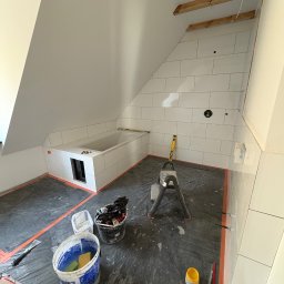 PRZEMYSŁAW DZIEDZIC PEPWO - Białe płytki ułożone w łazience na poddaszu, wanna obudowana płytkami, drabina i narzędzia glazurnicze widoczne na folii ochronnej.