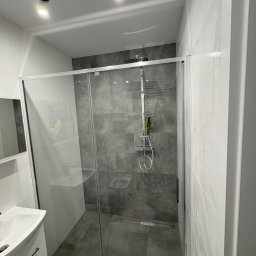 PRZEMYSŁAW DZIEDZIC PEPWO - Nowoczesna kabina prysznicowa z szarymi płytkami, biała umywalka z szafką, oświetlenie sufitowe. Minimalistyczny design łazienki po remoncie.