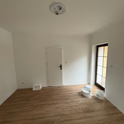 PRZEMYSŁAW DZIEDZIC PEPWO - Pusty pokój po remoncie z drewnianą podłogą, białymi ścianami z listwami i drzwiami. Na podłodze widoczne akcesoria malarskie.