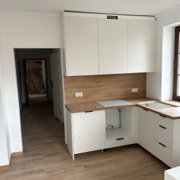 PRZEMYSŁAW DZIEDZIC PEPWO - Jasna kuchnia w trakcie montażu: białe szafki z drewnianymi blatami i panelami, brązowe płytki na podłodze, widoczne gniazdka elektryczne i otwarte przejście do innego pomieszczenia.