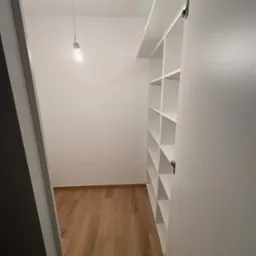 Wnętrze garderoby po remoncie: drewniana podłoga, białe ściany i regał z półkami. Widoczne oświetlenie w postaci wiszącej żarówki. Kątowe ujęcie z otwartymi drzwiami.